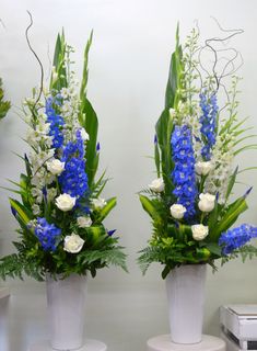 two white vases with blue flowers and greenery on the top are sitting side by side