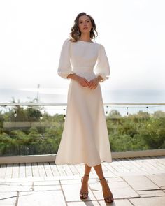 a woman wearing a white dress and heels standing on a brick floor with her hands in her pockets