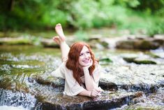 a woman laying on top of a rock next to a river with her arms in the air