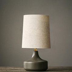 a gray table lamp with a white linen shade on the base and a light grey wall behind it