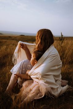 a woman holding a child wrapped in a blanket while sitting on the ground near some tall grass