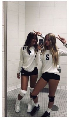 two women in white shirts and black shorts taking a selfie in a bathroom mirror