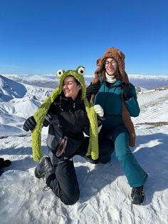 two people sitting in the snow with hats on and one holding a stuffed animal's head