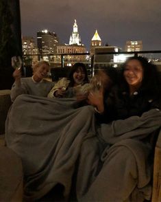 three women sitting on a couch covered in blankets and smiling at the camera with city lights behind them