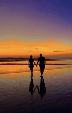 two people holding hands while walking on the beach at sunset or dawn with their reflection in the wet sand