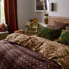 a bed with colorful sheets and pillows in a room next to a chair, table and window