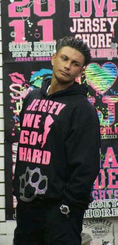 a young man standing in front of a wall with posters on it and wearing a black hoodie