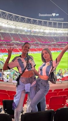 two women standing in the stands at a soccer game with their hands up and one holding her purse