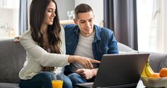 a man and woman sitting on a couch looking at a laptop screen with orange juice in front of them