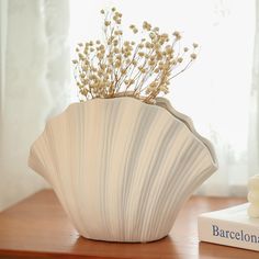 a white vase with flowers in it sitting on a table next to a book and window