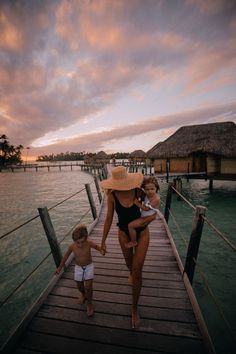 a woman and child walking on a dock in the water at sunset with huts in the background