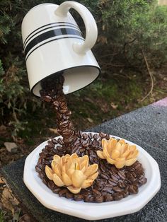 a coffee cup pouring water into a plate of coffee beans with flowers on top, in front of some trees