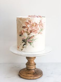 a white cake with pink flowers on it sitting on a wooden stand next to a wall