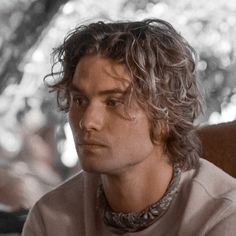 a man with long hair sitting in a chair looking off to the side while wearing a necklace