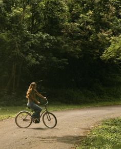 a woman riding a bike down a dirt road in the woods with trees behind her