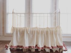 a white crib with pink ruffles in front of two windows and a wooden floor