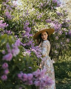 a woman in a dress and hat standing next to purple flowers with her hands on her hips