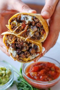 someone is holding two burritos with meat and cheese on them, in front of some guacamole