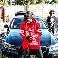 a man sitting on the hood of a car looking at his cell phone while another person stands next to him