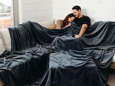 a man and woman sitting on a couch wrapped in black sheets with their arms around each other