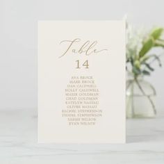 a white table number card with gold foil lettering on it and a vase full of flowers in the background