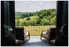 two chairs sitting on top of a balcony next to an open door that leads to a lush green field