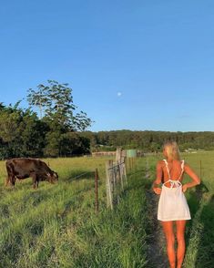 a woman in a white dress is walking down a path towards a cow grazing on the grass