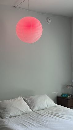 a bed with white sheets and pillows under a pink light hanging from the ceiling above it