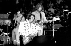 a man playing guitar while standing next to other men in front of microphones and an audience