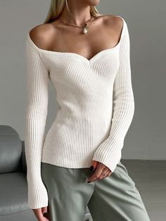 a woman wearing a white sweater and green pants posing in front of a gray couch