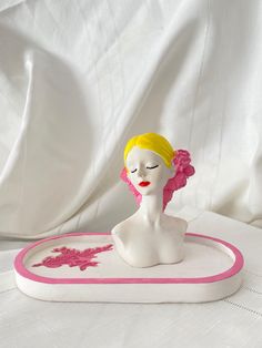 a white mannequin with pink trim and a flower in her hair sitting on a tray