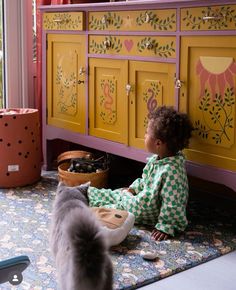 a small child sitting on the floor in front of a toy cabinet and cat looking at it