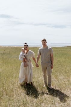 a man and woman holding hands walking through a field with a baby in her arms