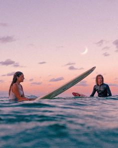 two women are sitting on surfboards in the ocean at sunset, one is holding her board