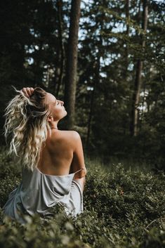 a woman sitting in the middle of a forest looking up into the sky with her hair blowing back