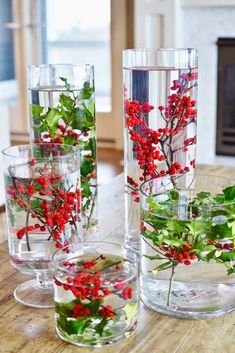 four glass vases filled with red berries and greenery on top of a wooden table