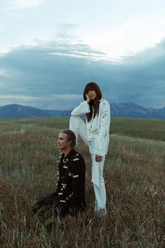 a man and woman sitting in the middle of a field with mountains in the background