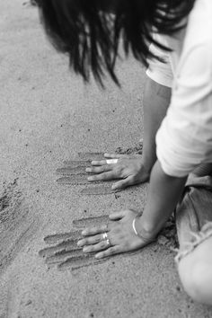 a woman kneeling down on top of a sandy beach next to her hand and footprints in the sand