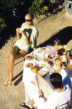 two women sitting at a table with food and drinks in front of them on the ground