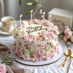 a birthday cake decorated with pink flowers and lit candles on a table next to goldware