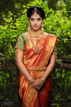 a woman in an orange and gold sari with jewelry on her neck, standing next to some bushes