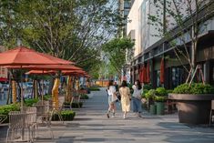 three women walking down the sidewalk in front of some shops and trees with red umbrellas