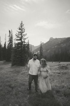 a man and woman are walking through the grass in front of some trees on a mountain