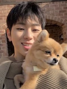 a young man holding a small dog in his arms and smiling at the camera while standing next to a brick wall