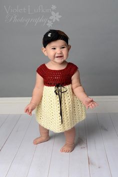 a smiling baby girl in a yellow and red dress standing on a white wooden floor