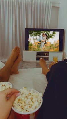 two people sitting on a couch watching tv and eating popcorn with their feet in the air