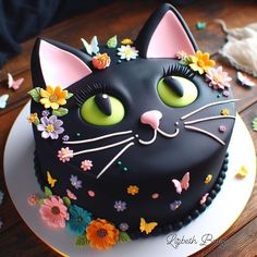 a black cat cake with flowers and butterflies on it's face is sitting on a plate