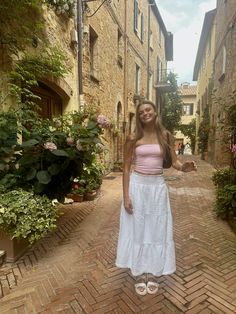 a woman standing in the middle of an alley way wearing a pink top and white skirt