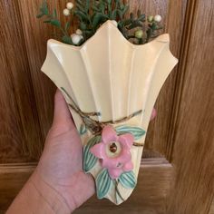 a hand holding a ceramic vase with flowers on the front and side, in front of a wooden door