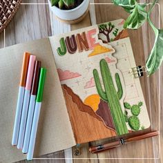 an open notebook sitting on top of a wooden table next to some pens and a potted plant